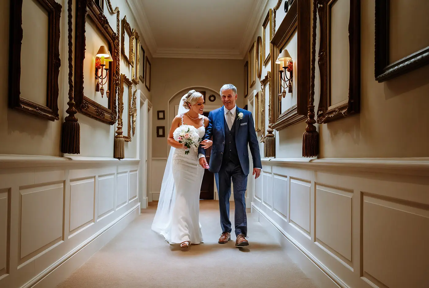 Bride Sally leaving her preparations room arm in arm with her father walking along a corridor of empty picture frames at Stubton Hall, Nottinghamshire
