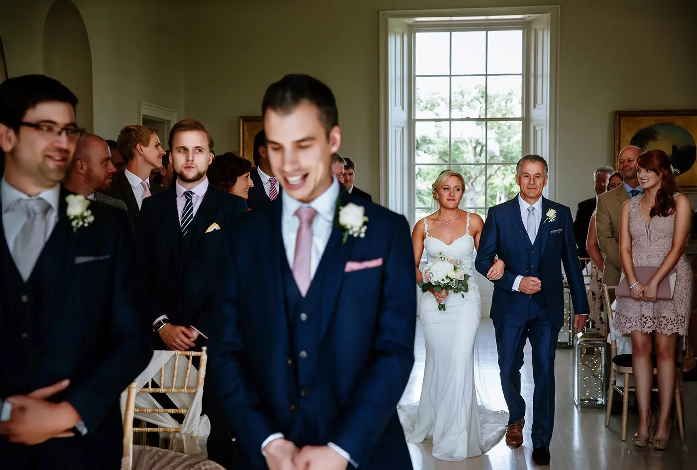 Bride Sally entering the ceremony room and walking the aisle with her father while groom Luke stands with his back to them at Stubton Hall, Nottinghamshire