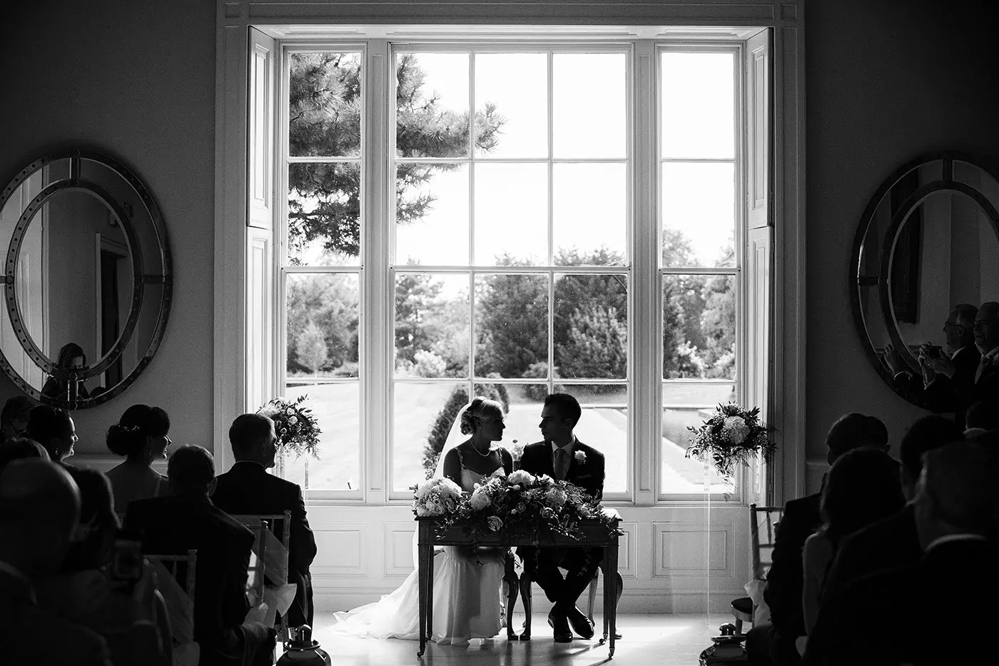 Black and white silhouette of bride Sally and groom Luke seated at the signing table with the grand window of Stubton Hall behind them
