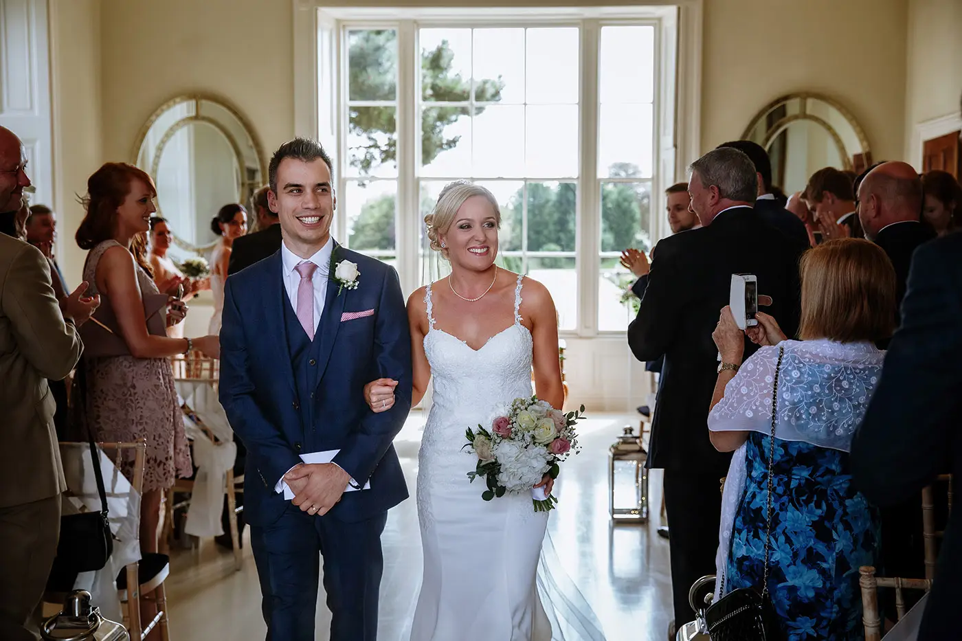 Bride Sally and groom Luke walking the recessional together smiling at their guests after the ceremony at Stubton Hall, Nottinghamshire