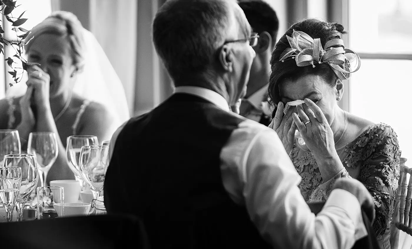 Mother of the groom crying and dabbing her tears with a tissue during the speeches at Stubton Hall while bride Sally looks on in the background